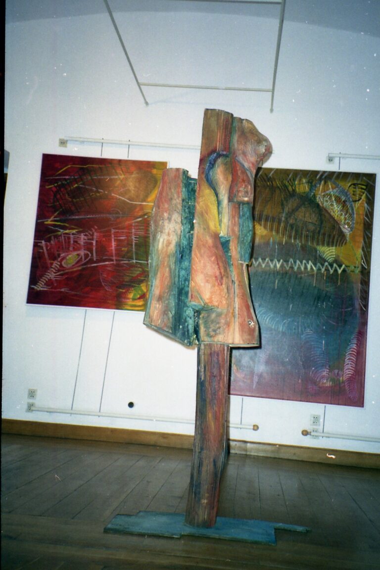 Tall painted sculpture standing before large gallery paintings